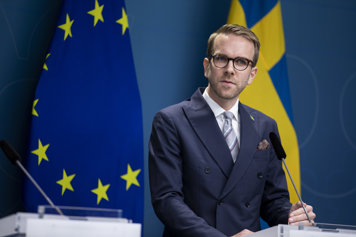 En person klädd i kostym står vid en talarpulpet framför en svensk och en EU-flagga.