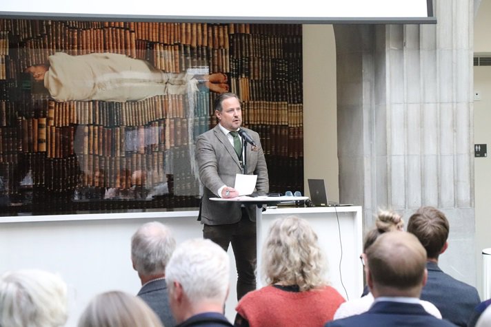 En man i kostym står och pratar framför en publik.