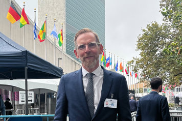 Socialministern står framför FN-skrapan i New York. Bakom honom syns en skyskrapa och en mängd flaggor på rad.