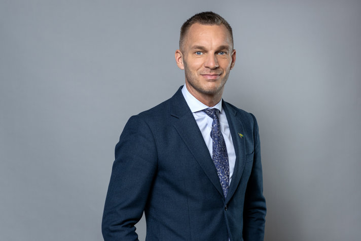 En person står framför en grå bakgrund. Personen är klädd i en mörkblå kostym, ljussblå skjorta och blå, blommig slips.