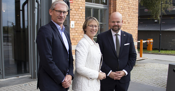 Tre personer tittar in i kameran och ler framför en tegelbyggnad.