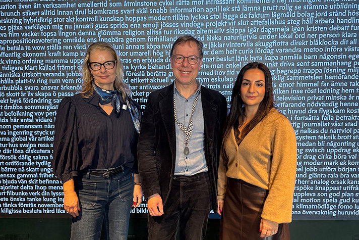 Lotta Edholm, Patrik Hadenius och Parisa Liljestrand står framför en blå vägg full med vit text.