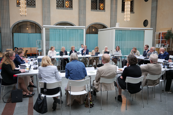 Ett tjugotal personer sitter längs fyra långbord som är vända mot varandra. Mötet hålls i en gammaldags banksal med valv och höga fönster.