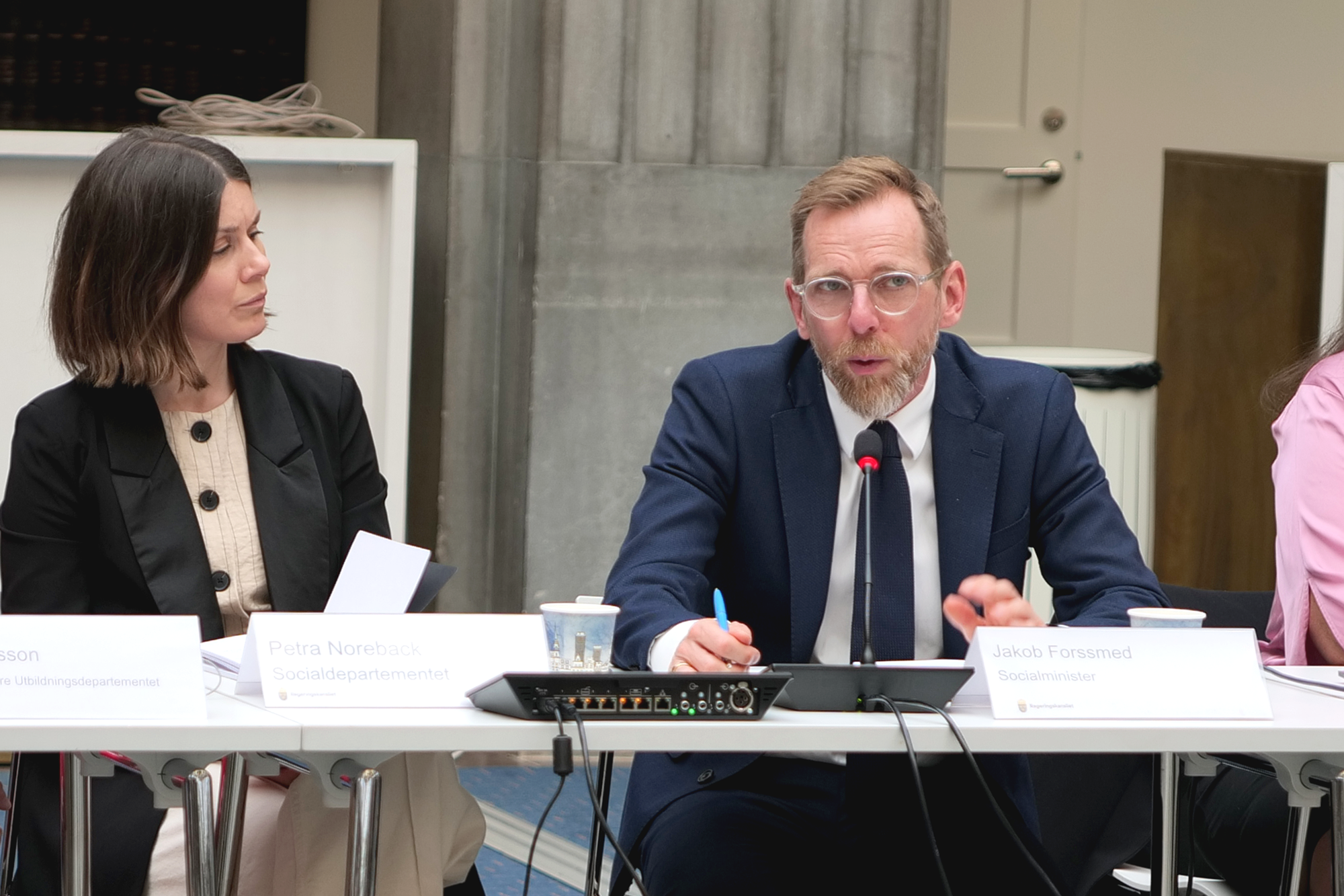 Socialminister Jakob Forssmed sitter vid ett bord och talar. Bredvid honom sitter statssekreterare Petra Noreback.