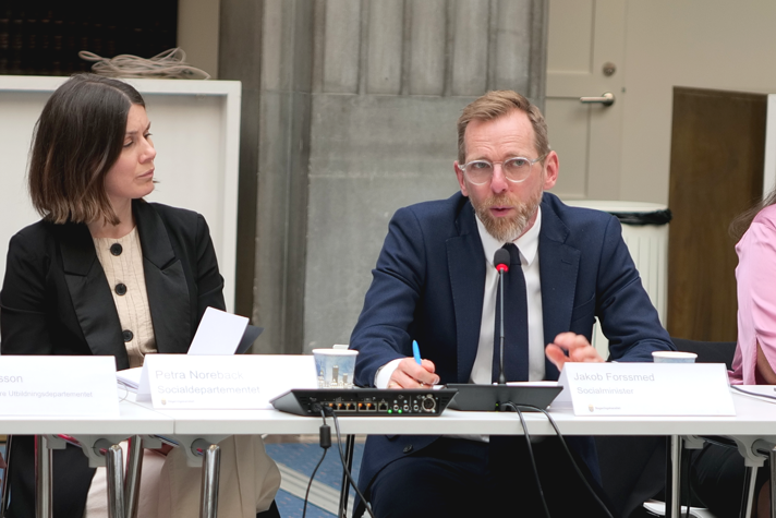 Socialminister Jakob Forssmed sitter vid ett bord och talar. Bredvid honom sitter statssekreterare Petra Noreback.