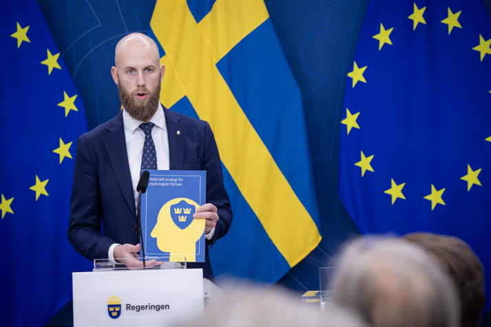 Carl-Oskar Bohlin står i regeirngens pressrum vid en talarstol och talar till journalister. I hand hand håller han en startegi där det stå på framsidan "Nationell strategi för psykologiskt försvar".
