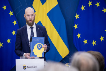 Carl-Oskar Bohlin står i regeirngens pressrum vid en talarstol och talar till journalister. I hand hand håller han en startegi där det stå på framsidan "Nationell strategi för psykologiskt försvar".