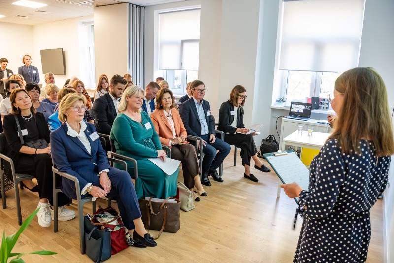 Socialtjänstministern och H.M. Drottningen tillsammans med andra mötesdeltagare lyssnar till en presentation. 