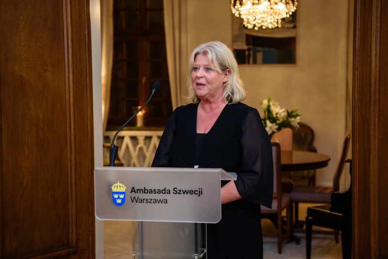 Socialtjänstministern står i en talarstol på ambassaden i Warszawa och håller ett anförande.