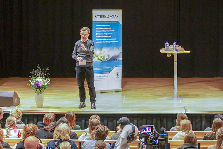 Ulf Kristerson står på en inomhusscen och talar. I förgrunden syns en publik med gymnasieelever.