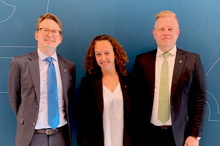 Tre personer står på rad framför en blå backdrop och ler mot kameran.