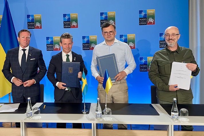 Bild på fyra personer som står bakom ett konferensbord. På skärmen bakom syns Natos logotyp och logotyp från Vilnius. 