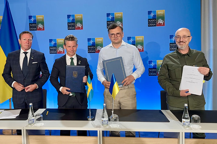 Bild på fyra personer som står bakom ett konferensbord. På skärmen bakom syns Natos logotyp och logotyp från Vilnius.