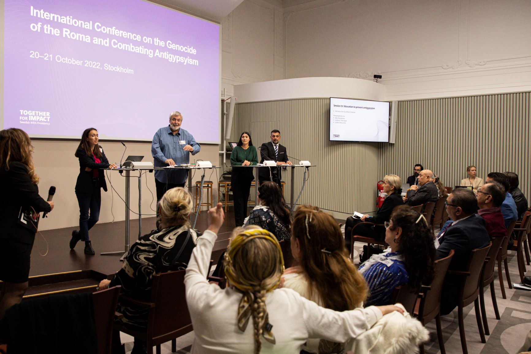 Bild från konferensen, talare på scenen och en i publiken gör gesten 'tummen upp'.