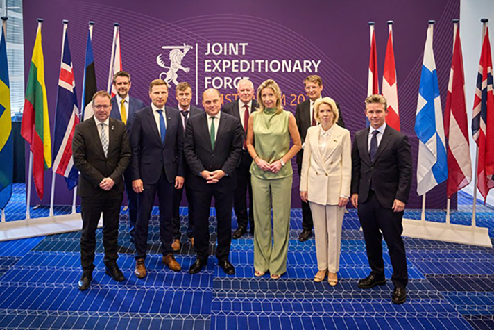 Gruppfoto på tio försvarsministrar som står framför tio flaggor. 