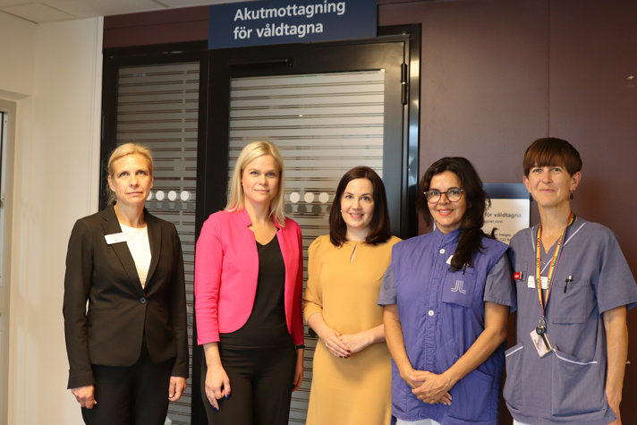 Fem personer, varav två vårdklädda personer, står i grupp framför en mottagningsdörr. På en skylt på väggen står det Akutmottagning för våldtagna