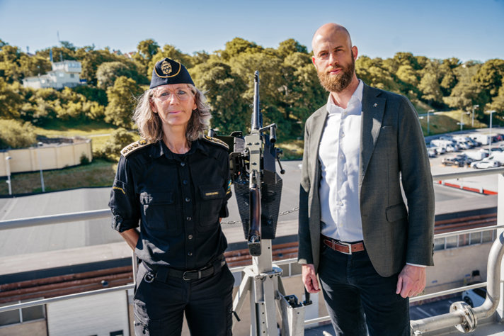 Lena Lindgren Schelin och Carl-Oskar Bohlin står på ett fartyg framför en kulspruta.