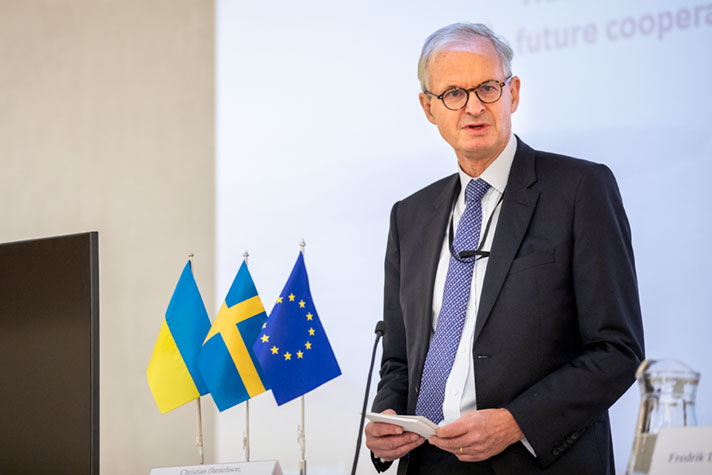 Christian Danielsson står vid ett talarbord på en scen inomhus. Ukrainska flaggan, svenska flaggan och EU-flaggan står på ett bord bredvid.