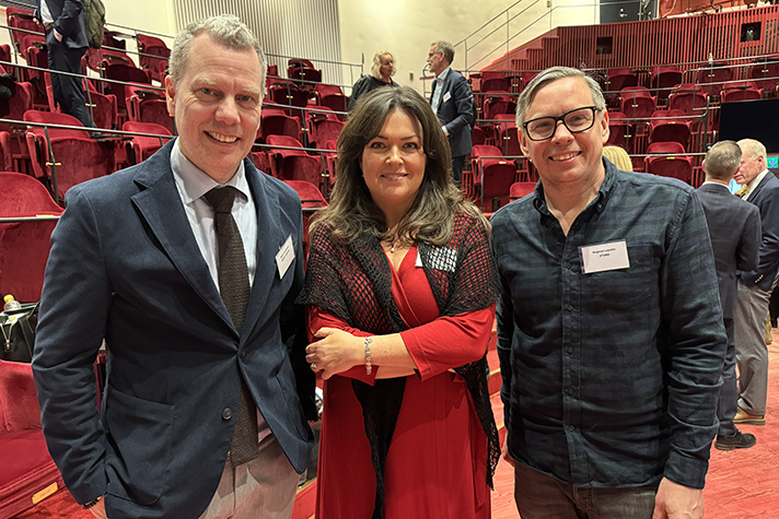 Tre personer fotograferas i halvfigur, stående i konferenslokalen.