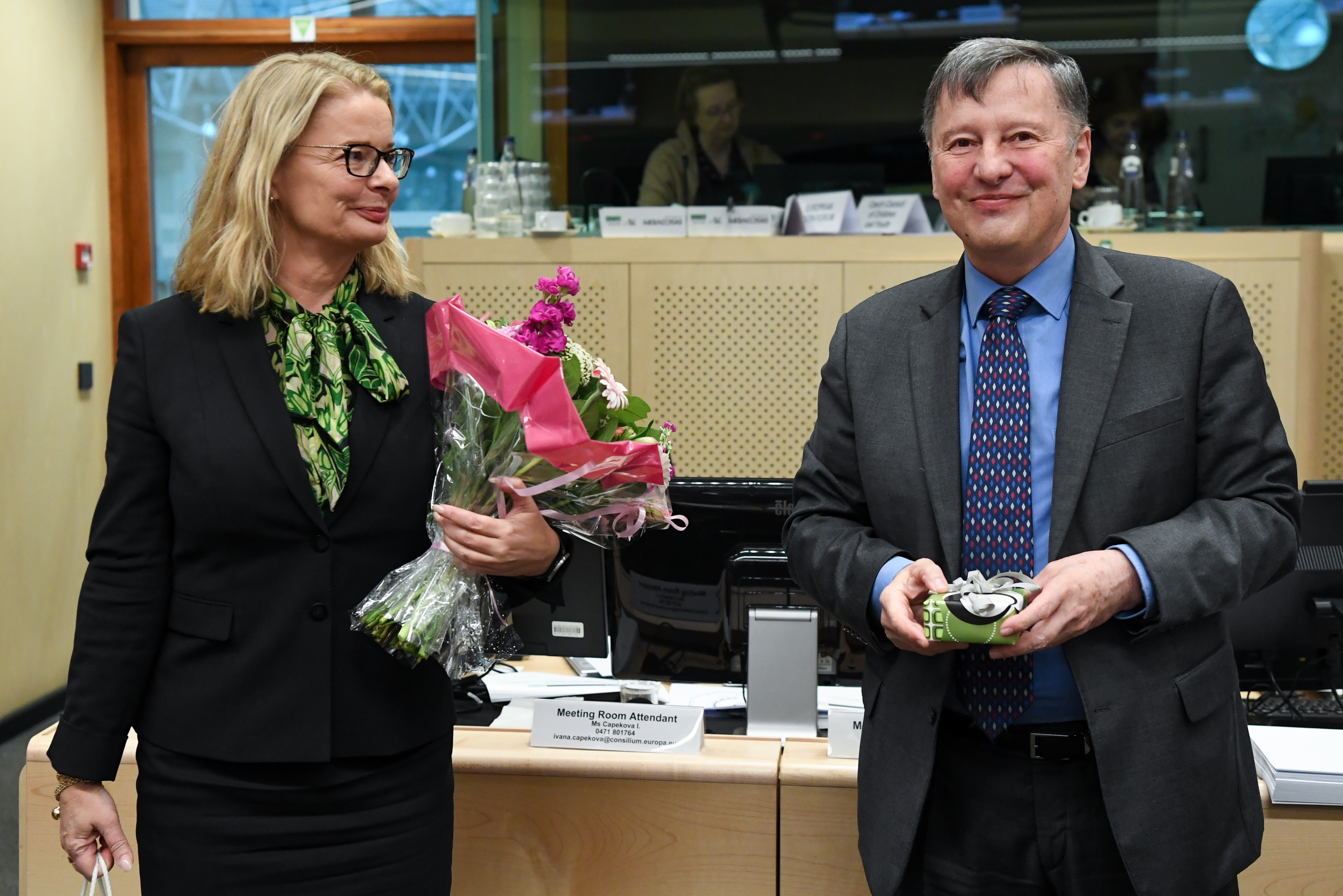 Två personer står bredvid varandra. Personen till vänster håller i en blombukett och tittar leendes på personen till höger. Personen till höger i bilden håller i en liten ask, tittar in i kameran och ler.
