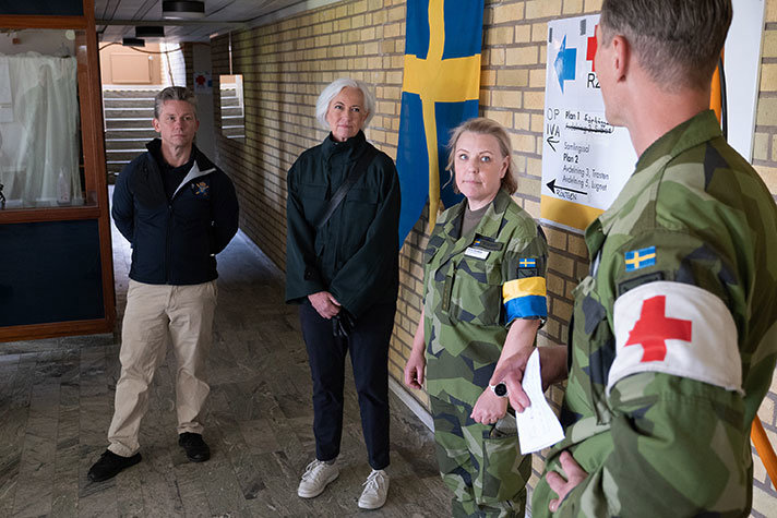 De två ministrarna stående inomhus med två sjukvårdare i militärkläder och svenska flaggan i bakgrunden.
