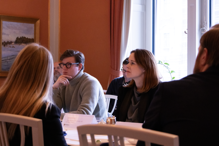 Statssekreterare Sara Modig och David Sikström sitter vid ett bord med namnskyltar. Runtomkring sitter resten av deltagarna vid rundabordssamtalet.