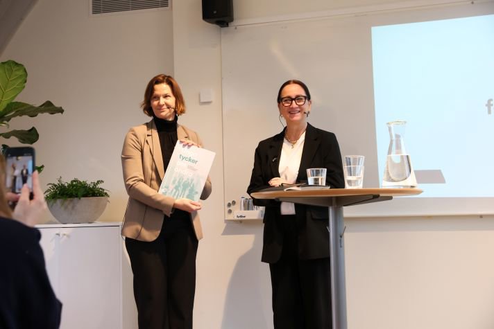 Bakom ett ståbord lämnar Petra Mårselius över en rapport till jämställdhetsminister Nina Larsson. I bakgrunden syns en grön växt och en whitboard. Rummet är ljust.