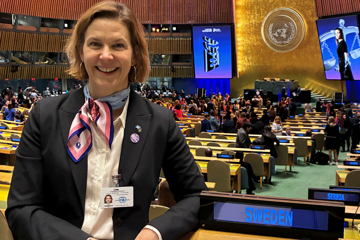 Nina Larsson står i ett FN:s stora mötesrum sidan om en skylt det står SWEDEN på. I bakgrunden syns långa bänkrader och stolar. En stor grupp människor står i mitten av rummet. Väggarna har träpaneler och bakom talarstolen längst bort i mötesrummet är en del av väggen förgylld med FN:s emblem på.
