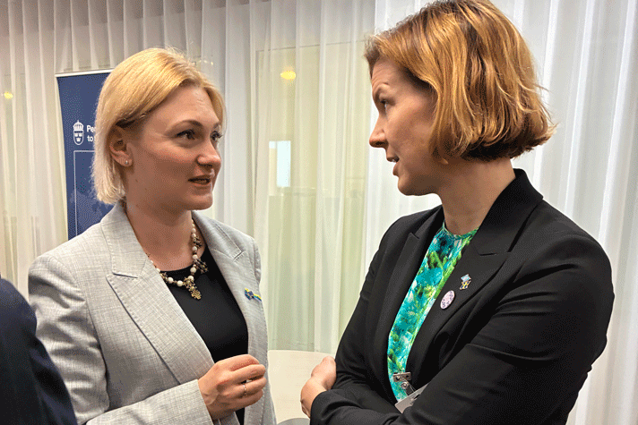 Parlamentsledamot Yevheniia Kravchuk från Ukraina och jämställdhetsminister Nina Larsson står och samtalar. I bakgrunden syns vita gardiner och en blå roll-up.