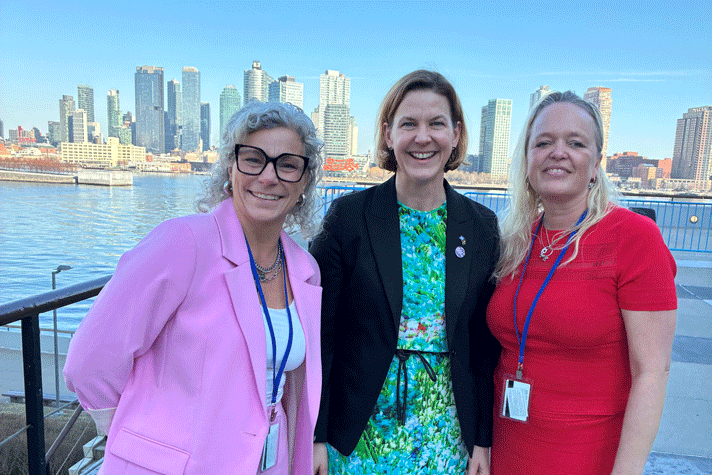 Åsa Johansen från Women in Tech, jämställdhetsminister Nina Larsson och Anna-Carin Hildingsson Boqvist från Ecpat står tillsammans framför havet i New York. En blå himmel och skyskrapor syns i bakgrunden syns