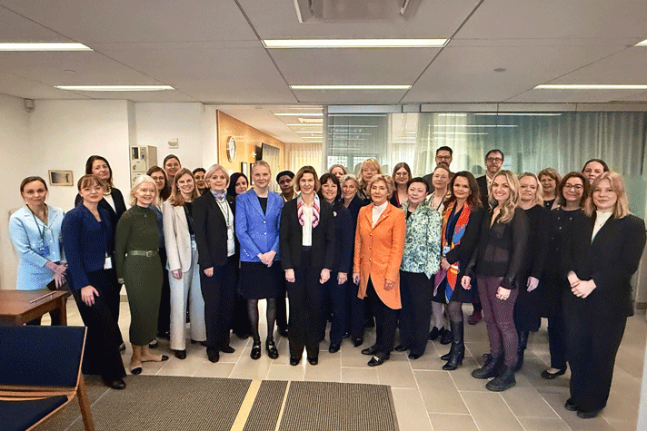 Hela den svenska delegationen med 30 personer står samlade i ett gruppfoto och ser in i kameran. I bakgrunden syns en glasvägg och en korridor. På golvet framför ligger en mörkgrå matta. Till vänster i bild syns några möbler.