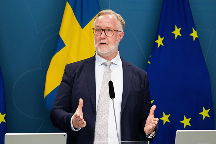 Arbetsmarknads- och integrationsminister Johan Pehrson talar på en pressträff. I bakgrunden syns en svensk flagga och en EU-flagga.