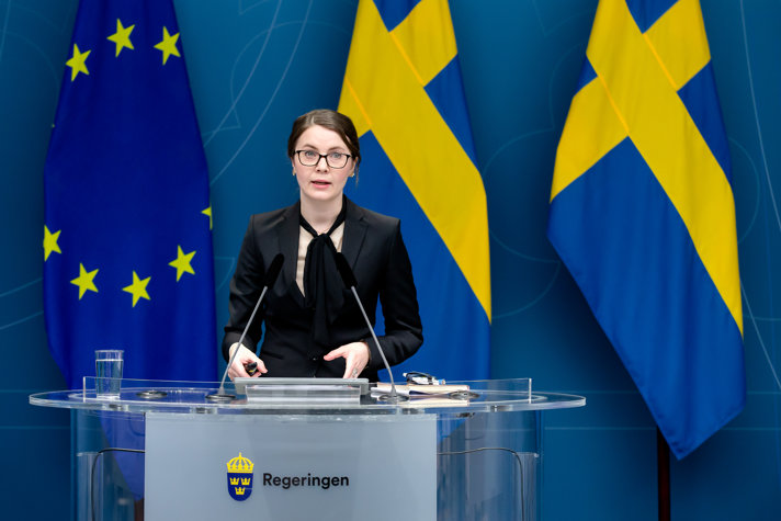 Ida Karkiainen står i en talarstol, i bakgrunden syns två svenska flaggor och EU-flaggan.