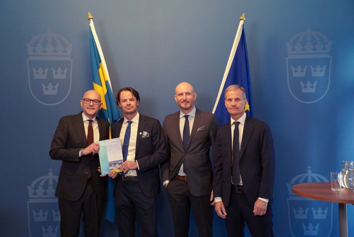 Ministern håller upp ansökan. De tre övriga står intill mot en bakgrund med RK:s logotyp.