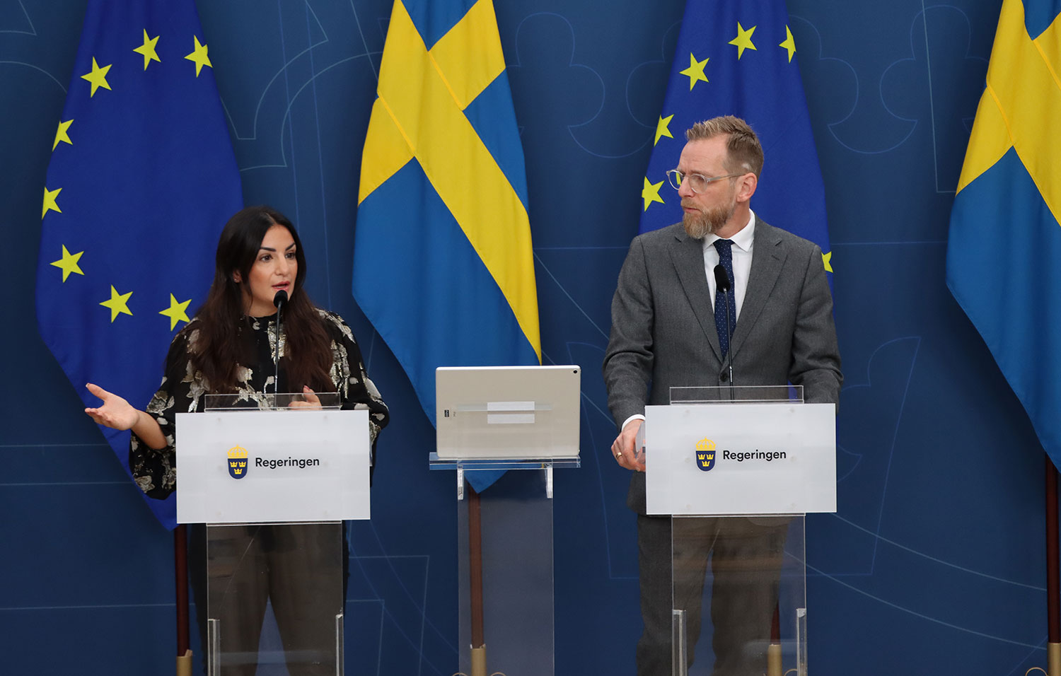 Parisa Liljestrand och Jakob Forssmed på pressträff. De står bakom varsitt prodium. Bakom dem en blå bakgrund och svenska och EU-flaggor.