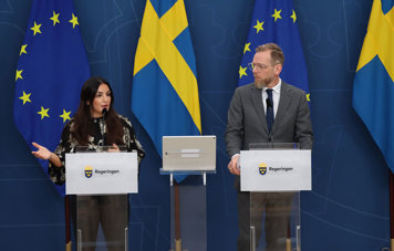 Parisa Liljestrand och Jakob Forssmed på pressträff. De står bakom varsitt prodium. Bakom dem en blå bakgrund och svenska och EU-flaggor.