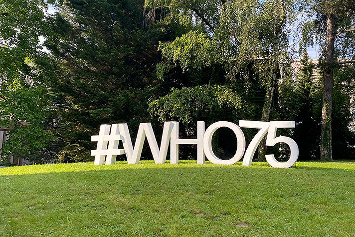 En gräsmatta med buskar i fonden. På gräsmattan står stora, vita tecken #WHO75 