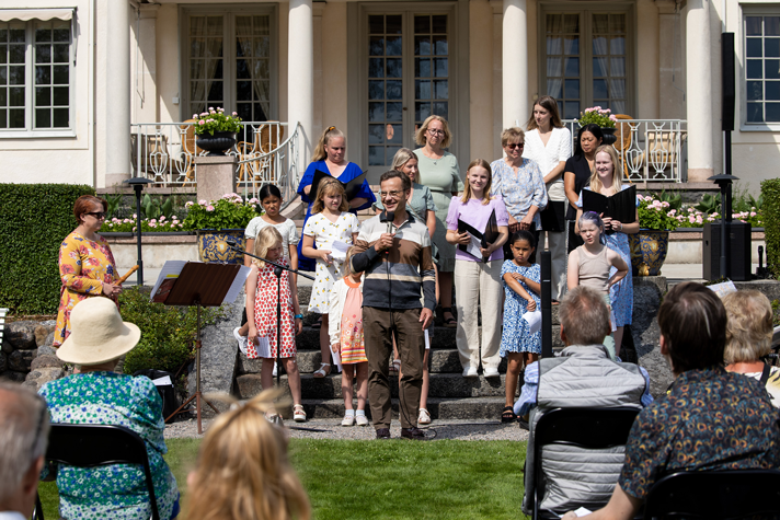 Statsministern håller i en mikrofon, sjungande barn runt omkring. 