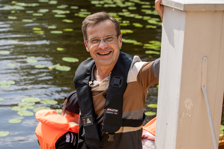 Statsministern står på en brygga, ett barn i flytväst i bakgrunden. 