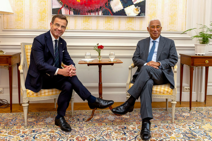 Ulf Kristersson och Antonio Costa sitter på varsin stol i äldre stil med ett litet runt bord mellan sig. På bordet står varsitt glas vatten. I bakgrunden syns en tavla och gulrandiga tapeter.