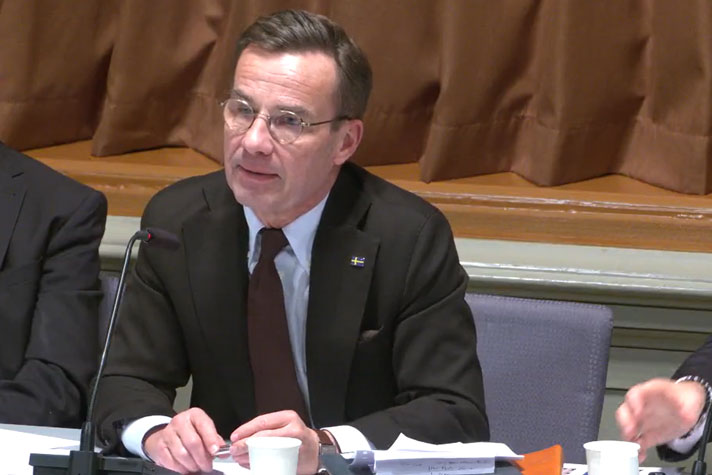 Statsministern sitter på en grå stol vid ett mötesbord. I bakgrunden syns ett brunt draperi.