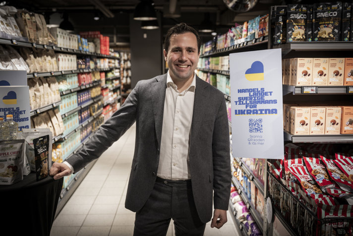Benjamin Dousa står i en matvarubutik bredvid en skylt med budskapet "Handelslandet Sverige tillsammans för Ukraina".
